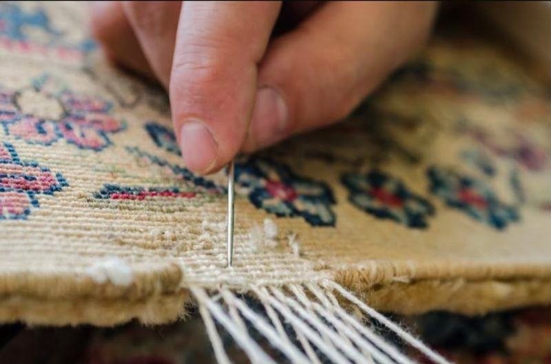 Main d’un lissier nouant une frange à l’aiguille sur un tapis crème